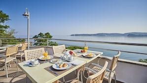 Un área de comedor al aire libre con una mesa preparada para el desayuno, con vistas a un paisaje escénico del mar y las colinas.