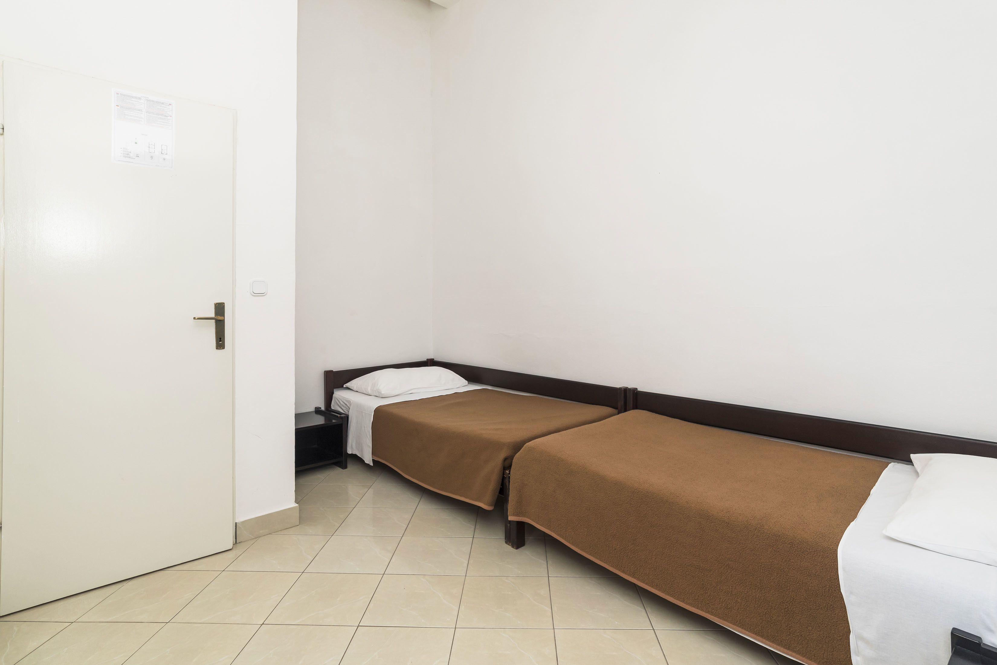 Habitació petita i neta amb dos llits individuals, una porta blanca i un terra de rajoles.