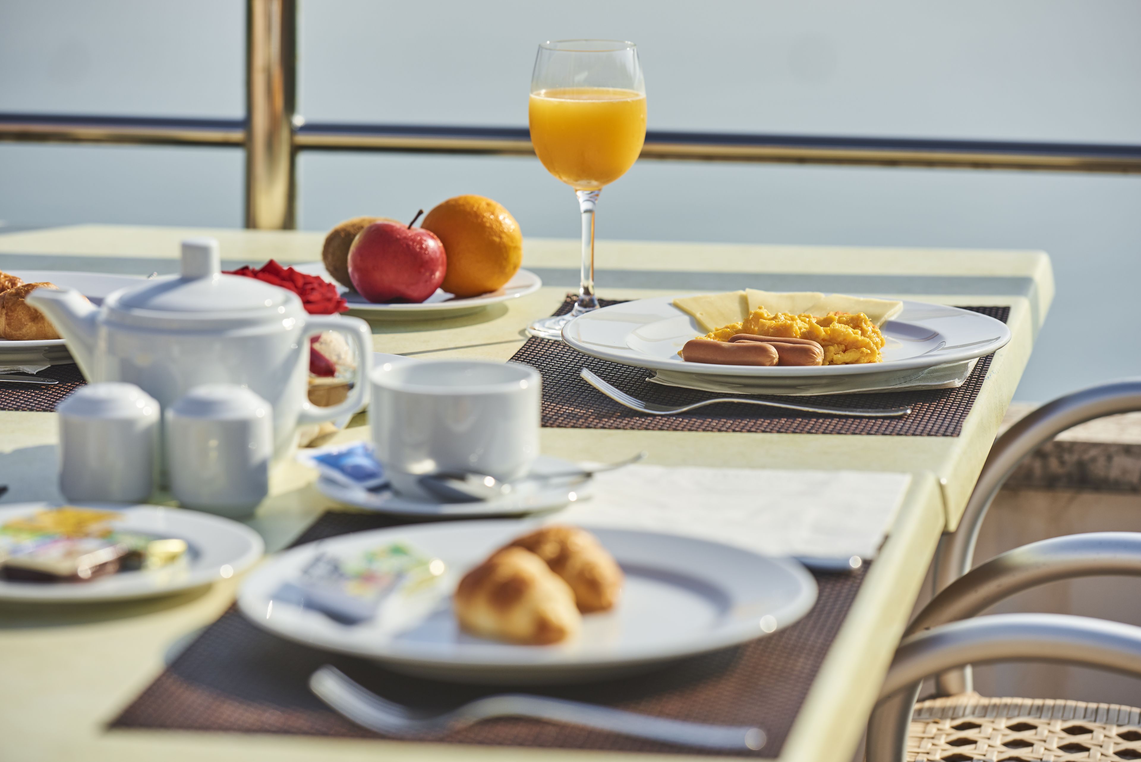 A breakfast table with various dishes, including croissants, fruit, and orange juice, set on a balcony with a view of the sea.