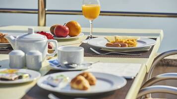A breakfast table with various dishes, including croissants, fruit, and orange juice, set on a balcony with a view of the sea.