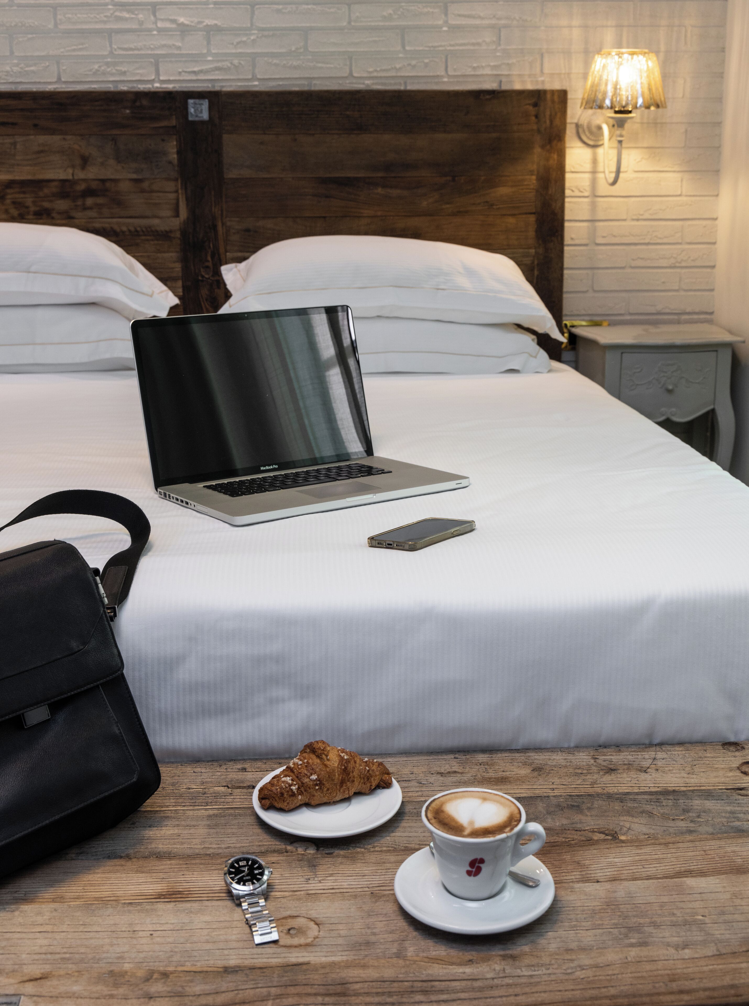A neatly made bed with a laptop, a smartphone, a croissant, a watch, and a cup of coffee on a wooden table.