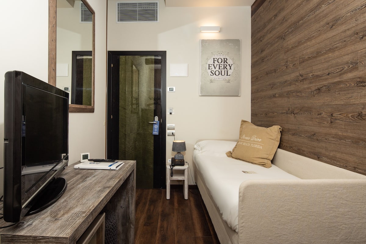 A hotel room with a wooden floor, a TV on a wooden table, a bed with a pillow, a small table with a lamp, and a wall with a 'For Every Soul' sign.