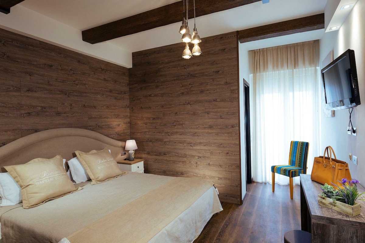 A bedroom with a wooden wall, a bed with beige bedding, a nightstand with a lamp, a chair, a TV, and a window with curtains.