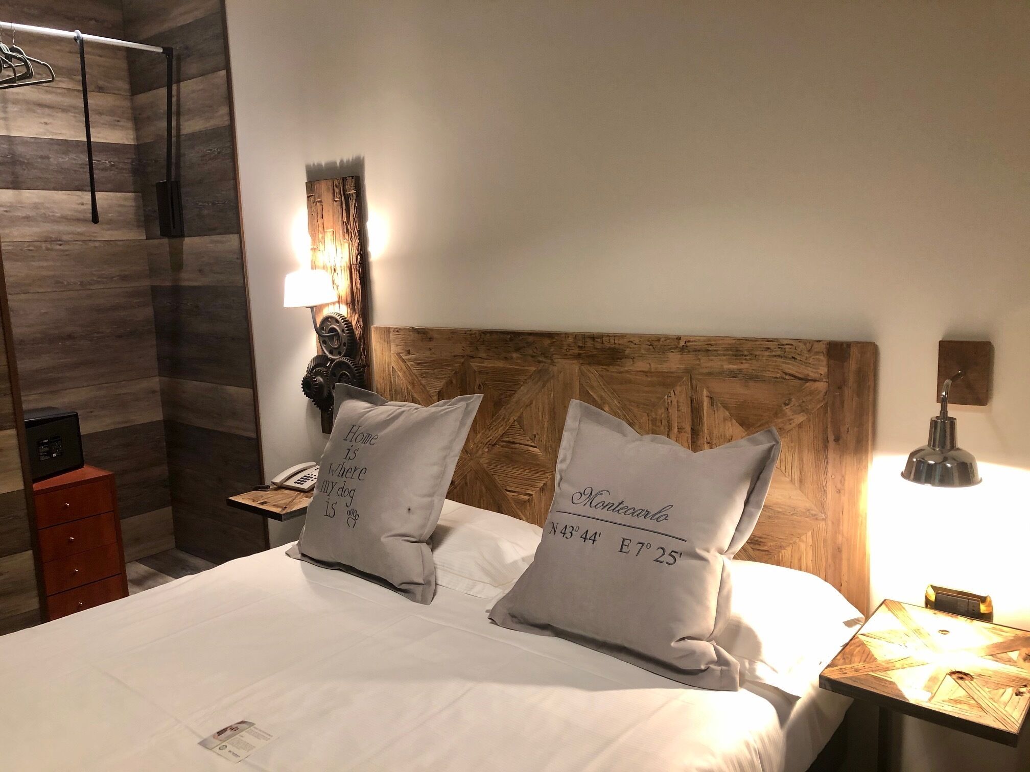 A neatly made bed with two pillows, a wooden headboard, a bedside lamp, and a small nightstand with a phone and a lamp.