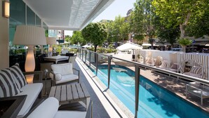 Ein Poolbereich mit Liegestühlen, einem Tisch und einem Glasgeländer mit Blick auf einen Swimmingpool.