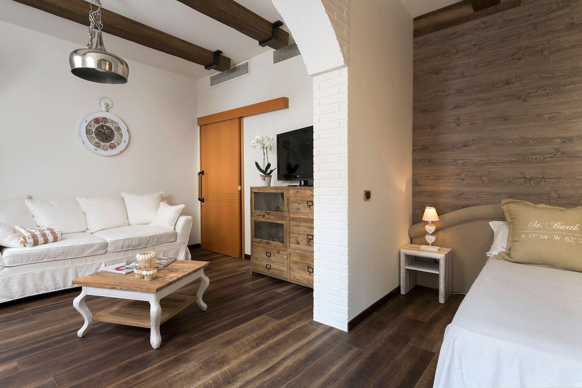 A modern hotel room with a white sofa, a wooden coffee table, a TV on a wooden cabinet, and a bed with a beige pillow.