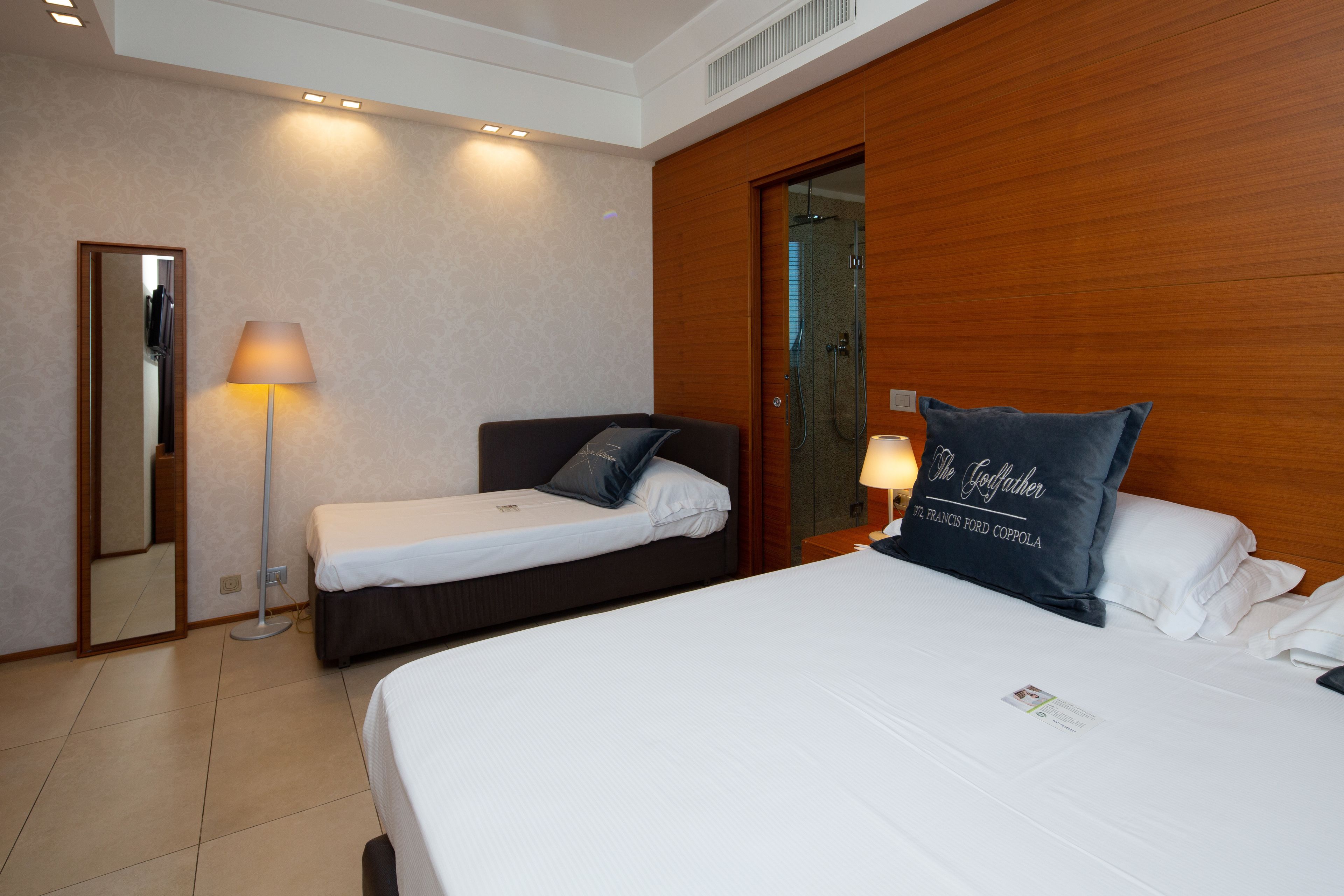 A hotel room with a large bed, a smaller bed, a mirror, a floor lamp, and a wooden wardrobe.