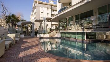 Un área de piscina moderna al aire libre con terraza de madera, sillas de descanso y una zona de estar cubierta de vidrio.