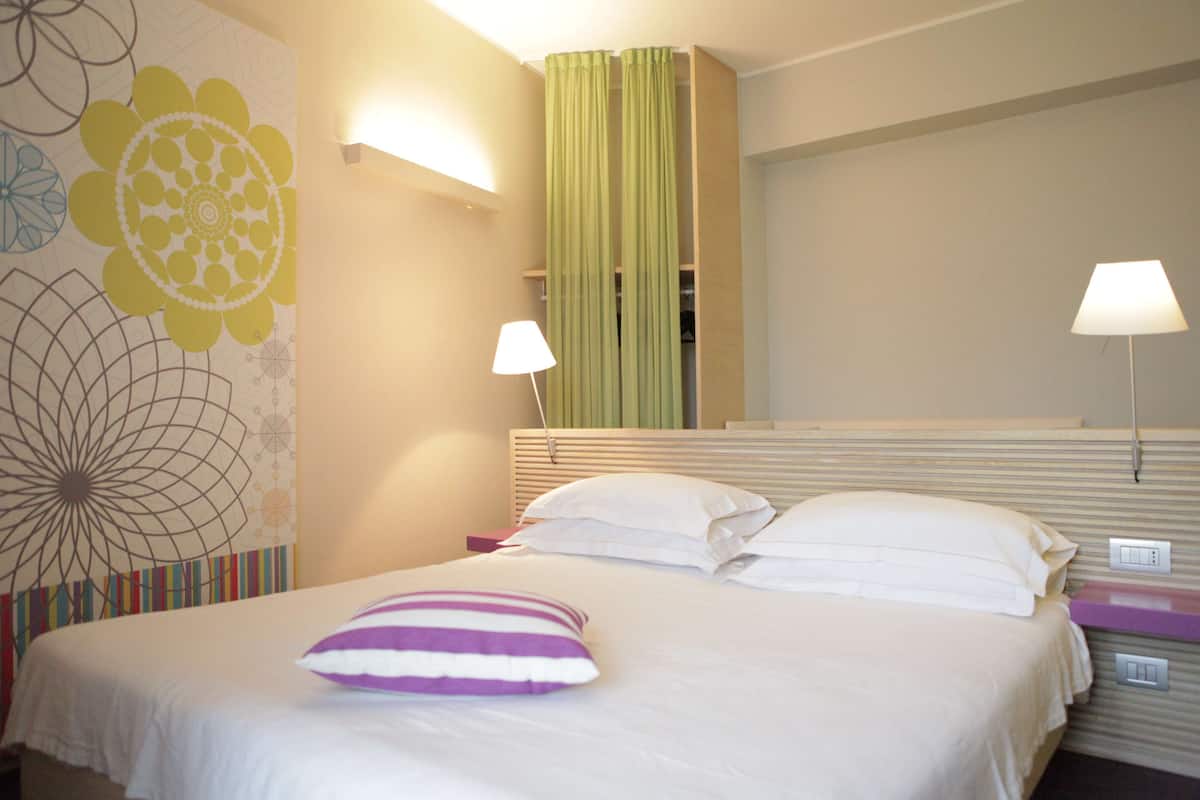 A hotel room with a bed, two bedside lamps, a wall with a floral design, and a green curtain.