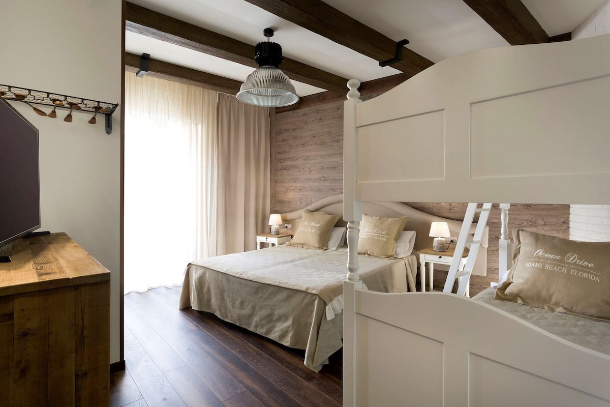 A bedroom with a wooden floor, a large bed with beige bedding, a nightstand with a lamp, and a ceiling fan.