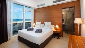 Ein modernes Hotelzimmer mit einem großen Bett, einer Glastür, die auf einen Balkon führt, einem Holz-Kopfteil und einem kleinen Schreibtisch mit einer Lampe.