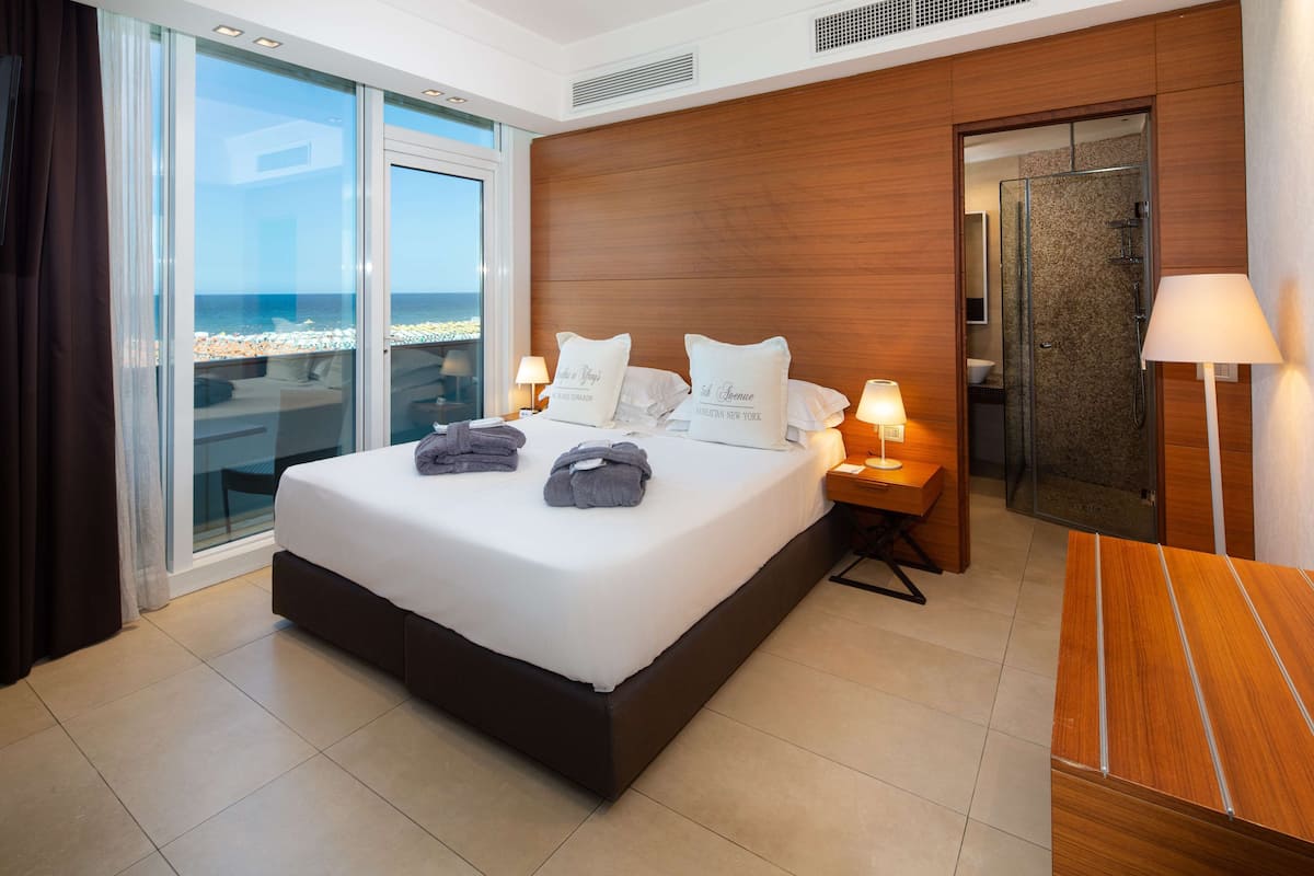 A modern hotel room with a large bed, a glass door leading to a balcony, a wooden headboard, and a small desk with a lamp.