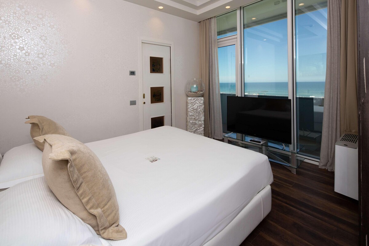 A hotel room with a large bed, a television, a glass door leading to a balcony with a view of the sea, and a small table with a glass top.