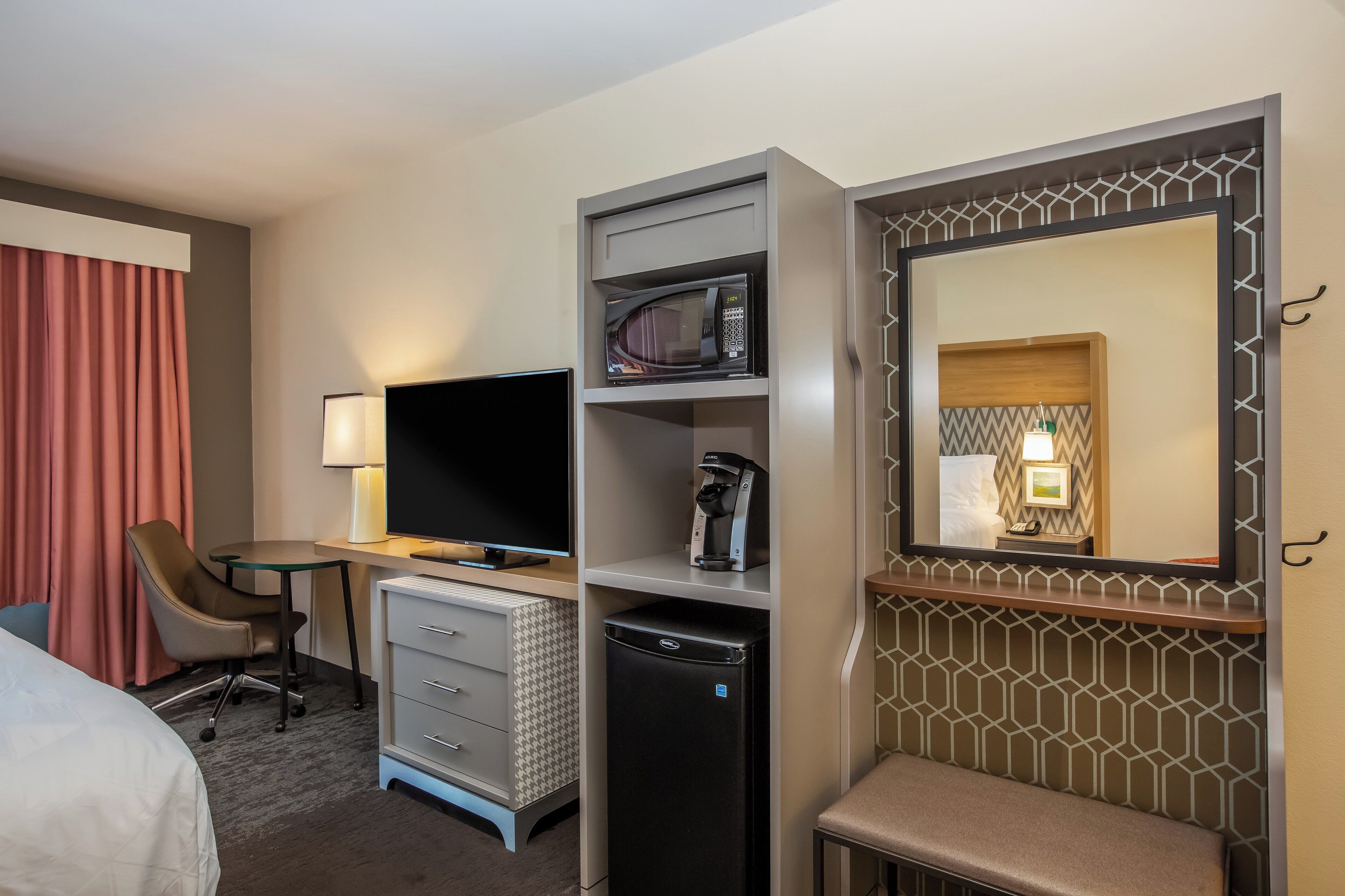 A hotel room with a bed, desk, chair, TV, microwave, coffee maker, and a mirror.