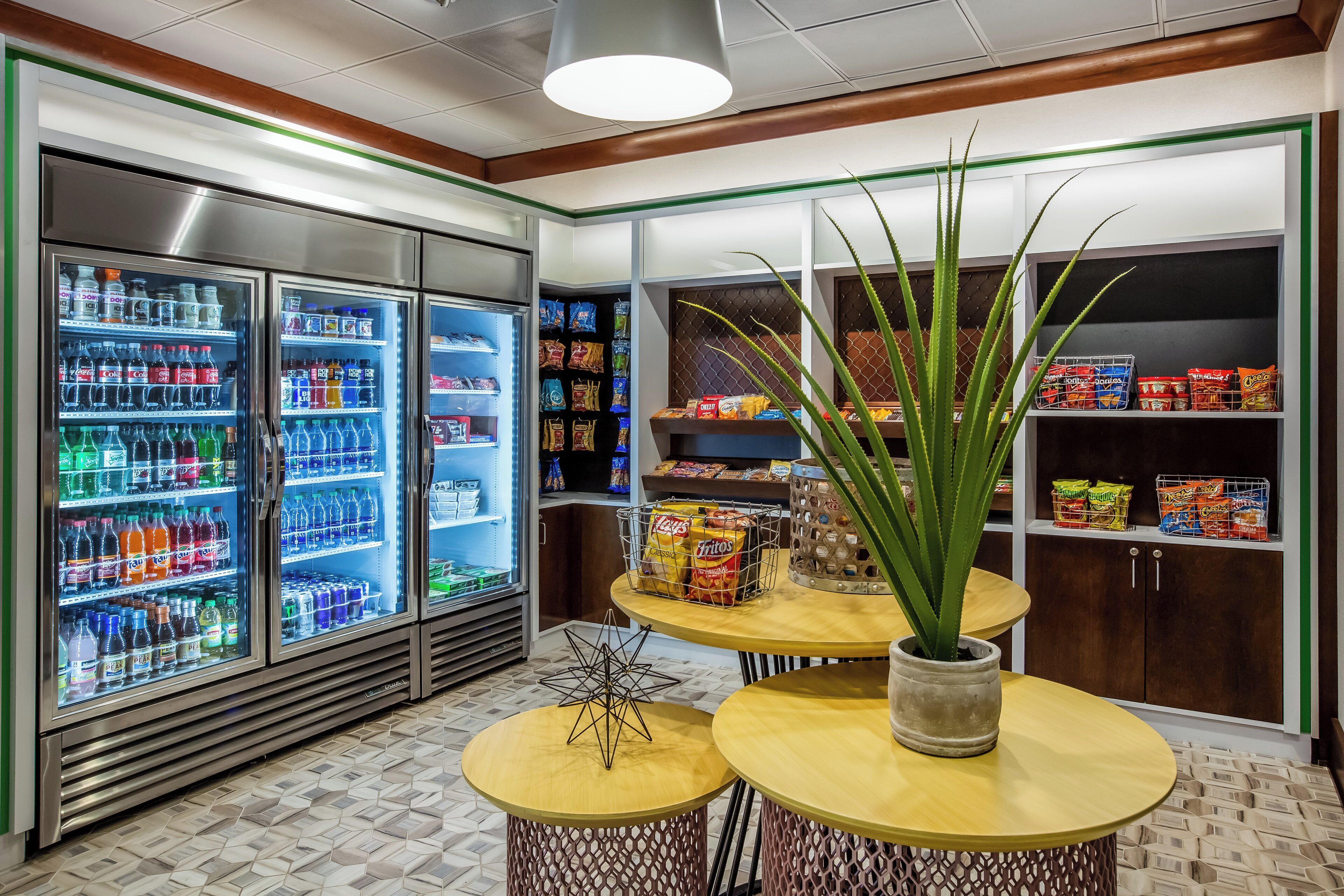 A snack bar area with refrigerated beverage dispensers, snack shelves, and a central table with a potted plant.