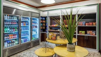 A snack bar area with refrigerated beverage dispensers, snack shelves, and a central table with a potted plant.