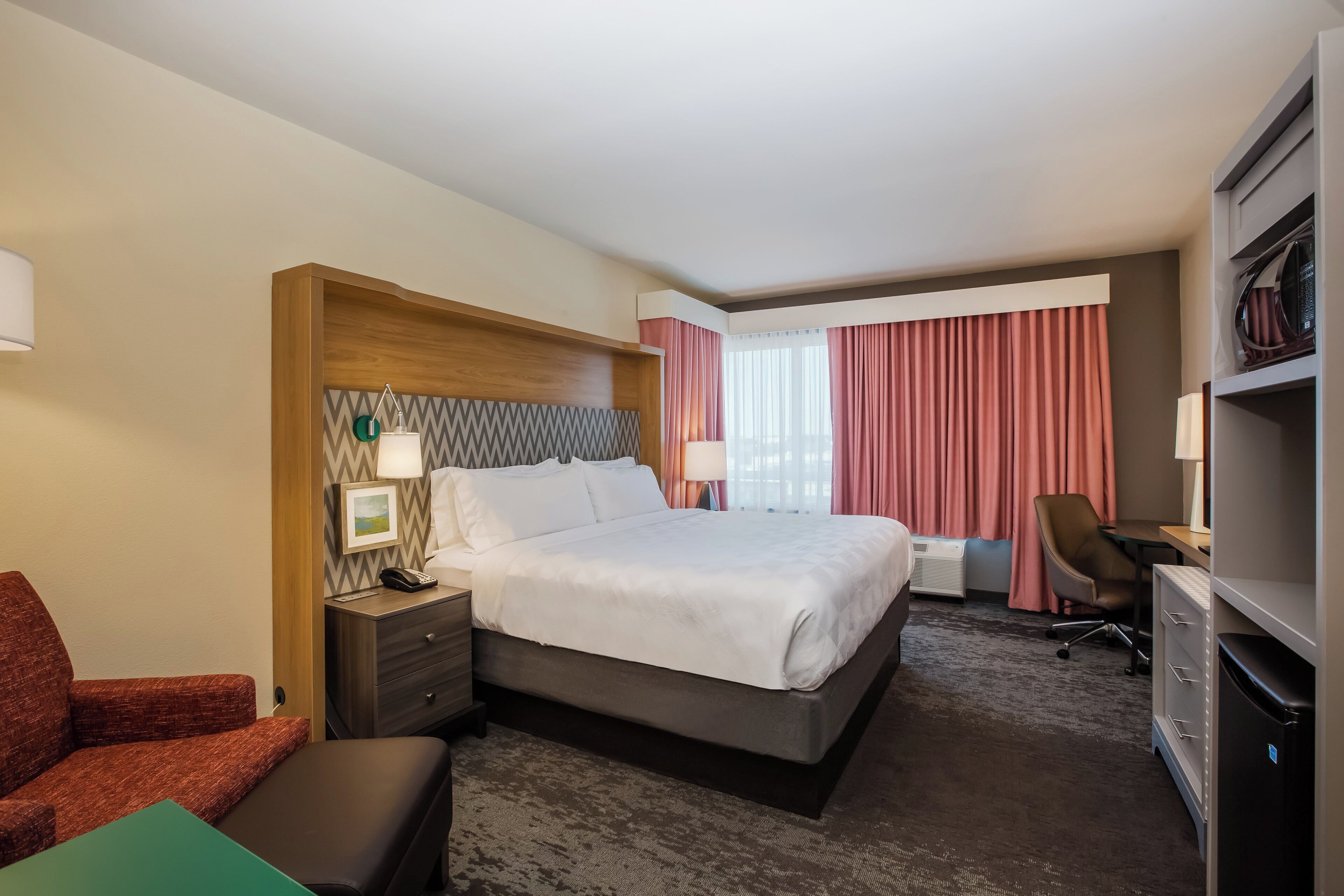 A hotel room with a bed, a desk, a chair, a TV, and a minibar.