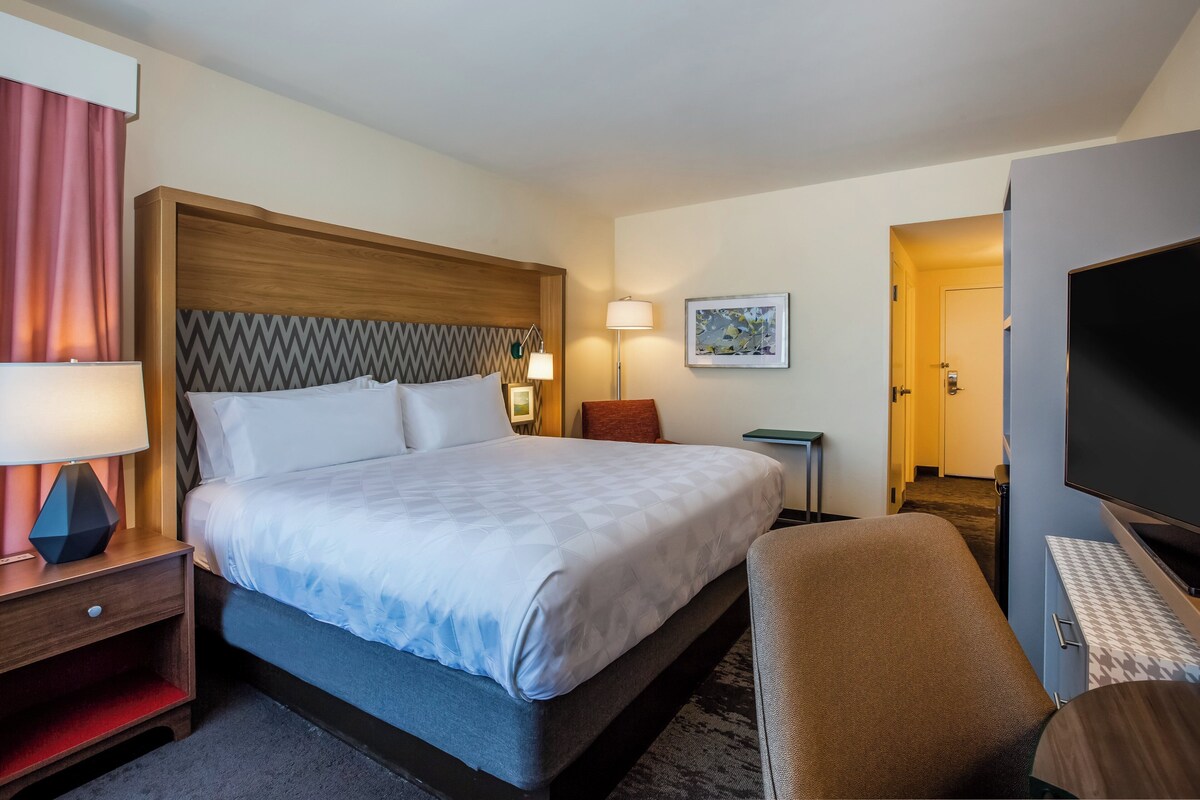 A hotel room with a bed, bedside table, lamp, TV, and a small desk.