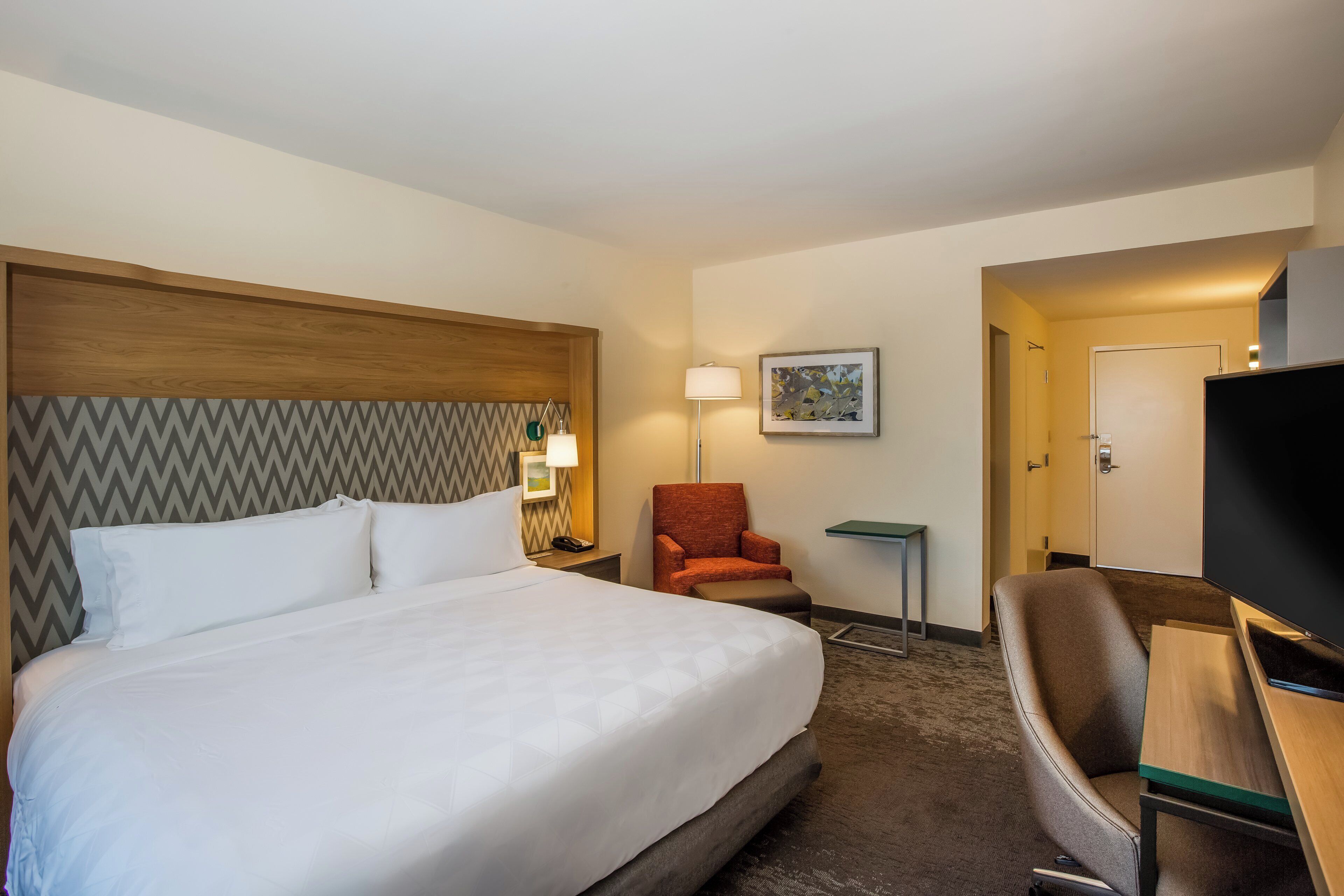 A hotel room with a bed, a chair, a desk, a lamp, and a television.