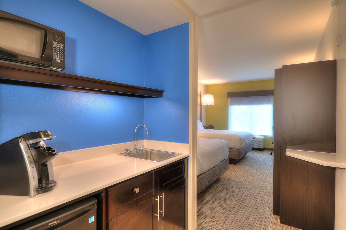 Une chambre d’hôtel équipée d’une kitchenette comprenant un four à micro-ondes, une machine à café et un évier. La chambre dispose d’un lit et d’une fenêtre avec des rideaux.