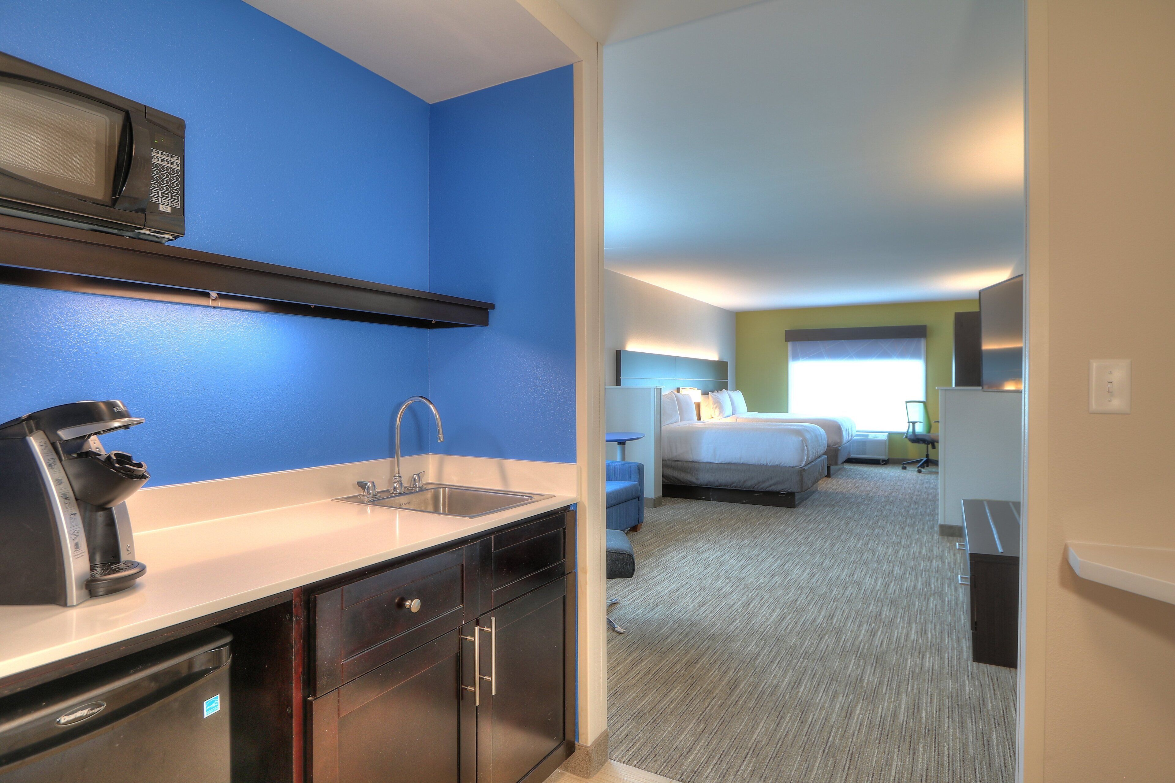 A hotel room with a kitchenette, a bed, a desk, and a bathroom.