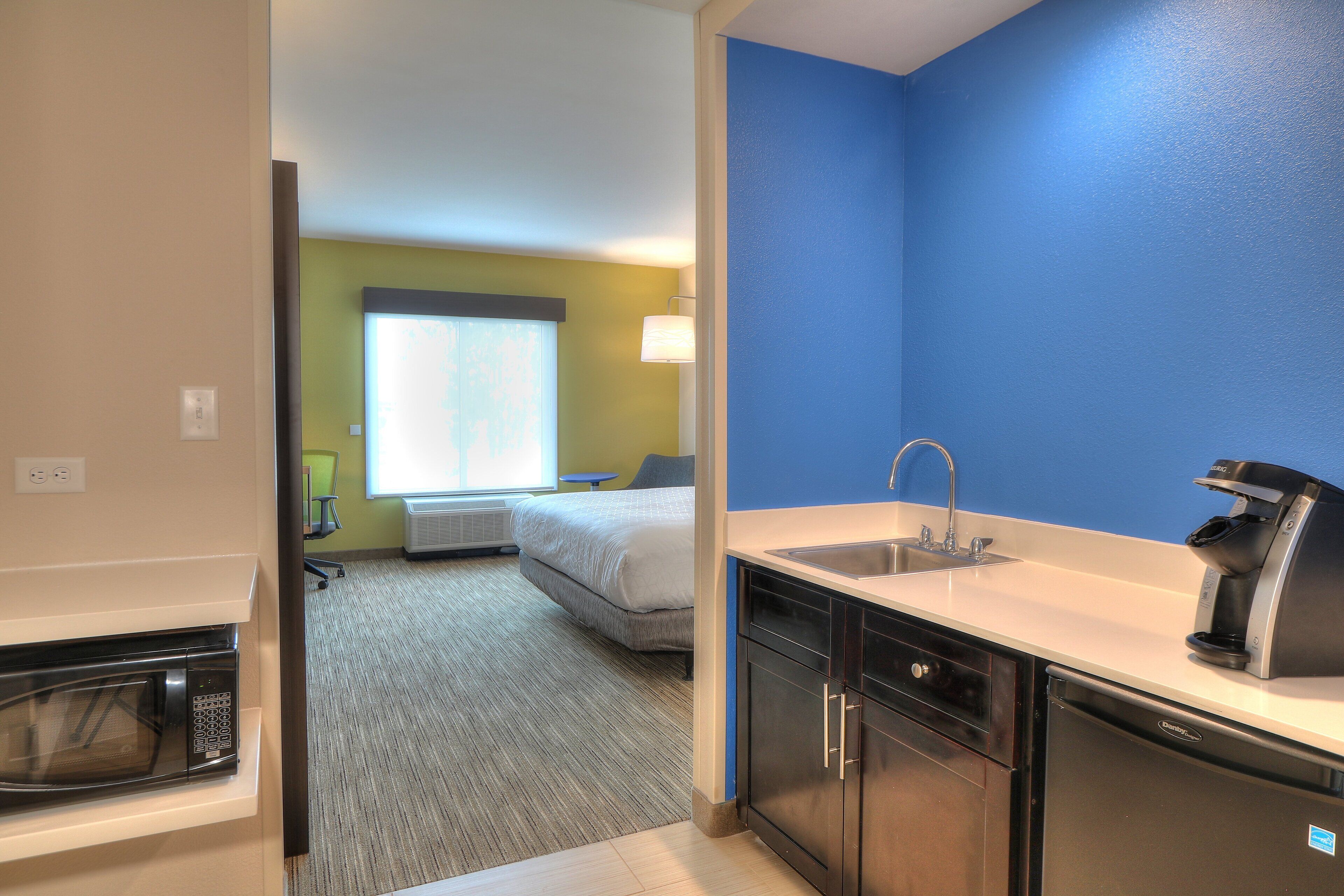 A compact hotel room with a kitchenette, a bed, a window with blinds, and a bathroom with a sink.