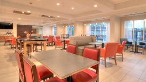 A modern dining area with wooden tables and red chairs, a sofa, and large windows with curtains. - Holiday Inn Express Hotel & Suites Mobile/Saraland by IHG (Saraland)