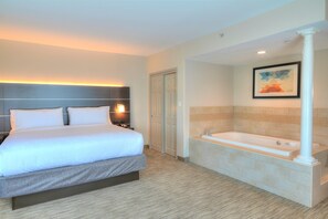 Habitación de hotel con cama, mesita de noche, lámpara, puerta y baño con bañera y toallero.