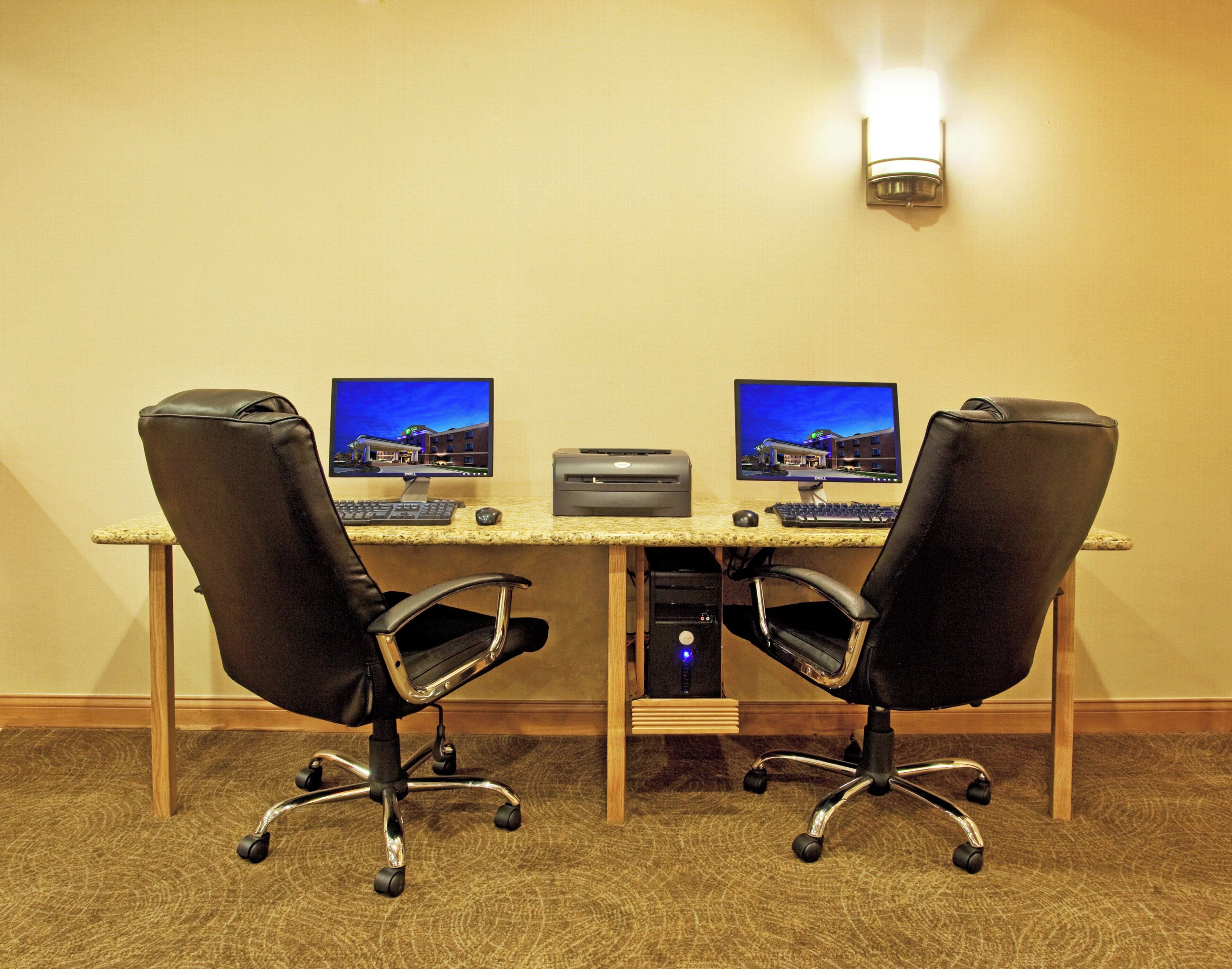 A hotel room with a desk, two chairs, two monitors, a printer, and a wall-mounted light fixture.