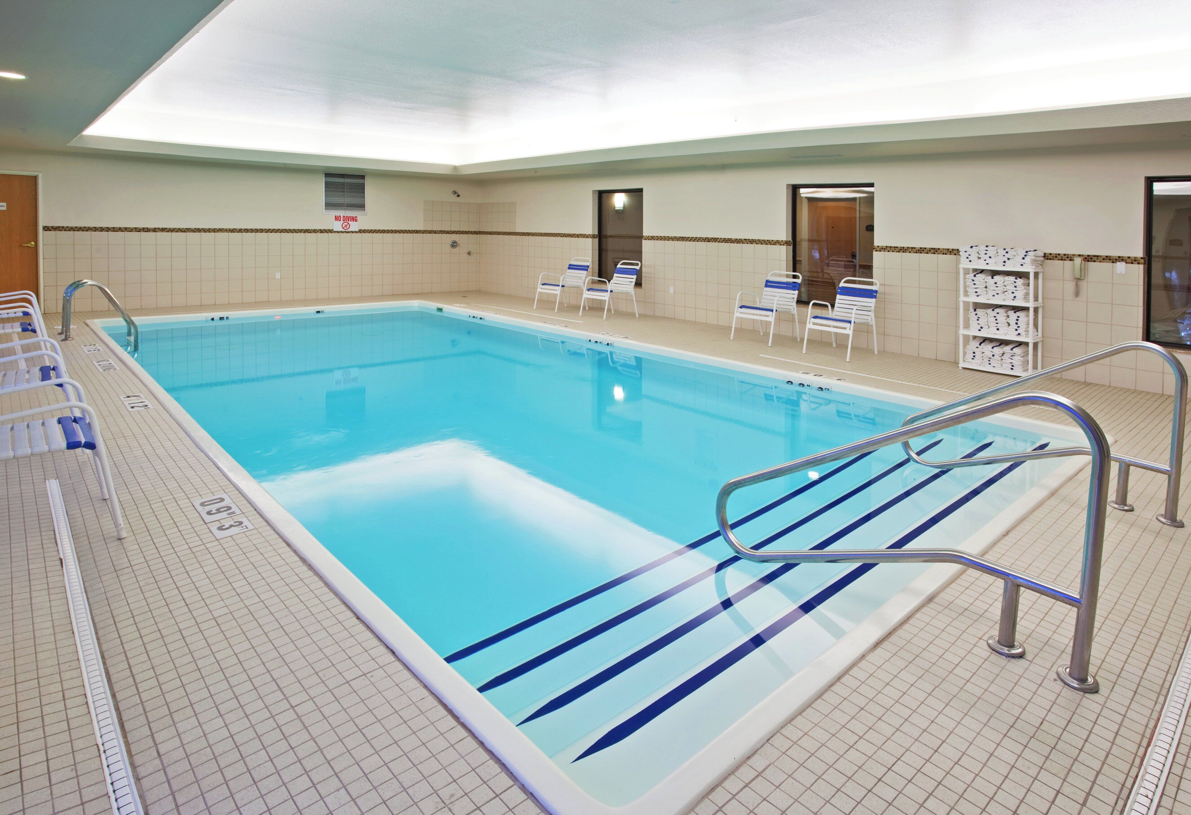 Una gran piscina interior con suelo de baldosas, un cartel de prohibición de saltos y varias sillas de descanso.