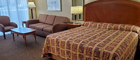 A hotel room with a large bed, a sofa, a small table, and a chair. There is a mirror on the wall and a window with curtains.
