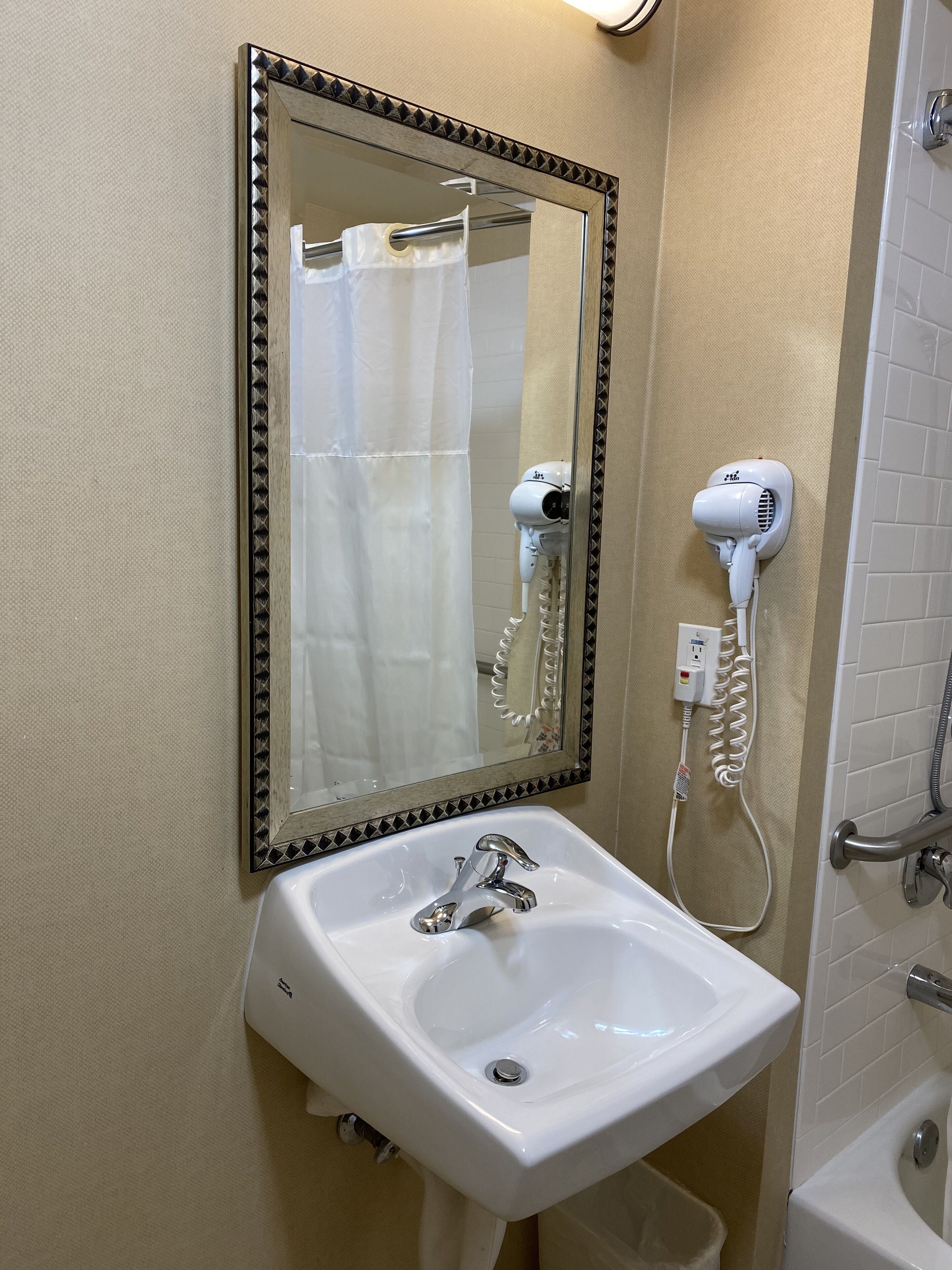 Un baño con lavamanos blanco, un espejo con marco decorativo, un secador de pelo y una zona de ducha.