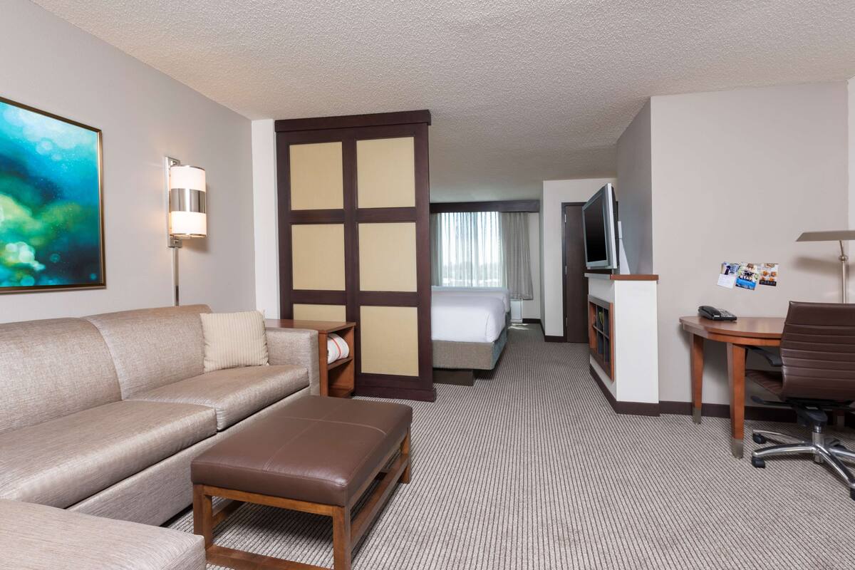 A hotel room with a sofa, a small table, a desk, a TV, a bed, and a large painting.