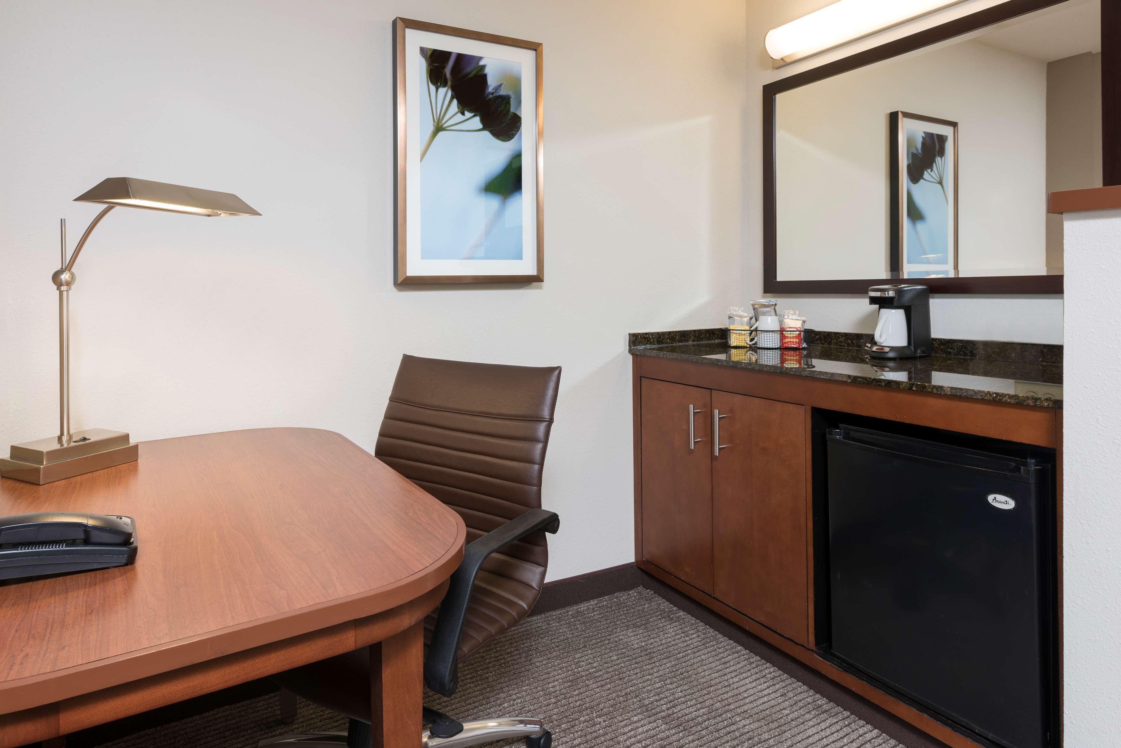 Une salle de conférence avec une table en bois, un fauteuil en cuir marron, une lampe de bureau, un réfrigérateur, une machine à café et des œuvres d’art encadrées accrochées au mur.