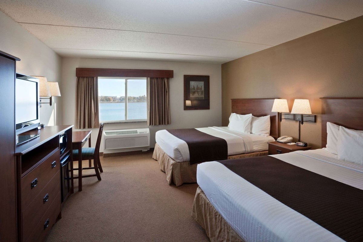 A hotel room with two beds, a desk, a chair, a TV, a window with curtains, and a view of the water.