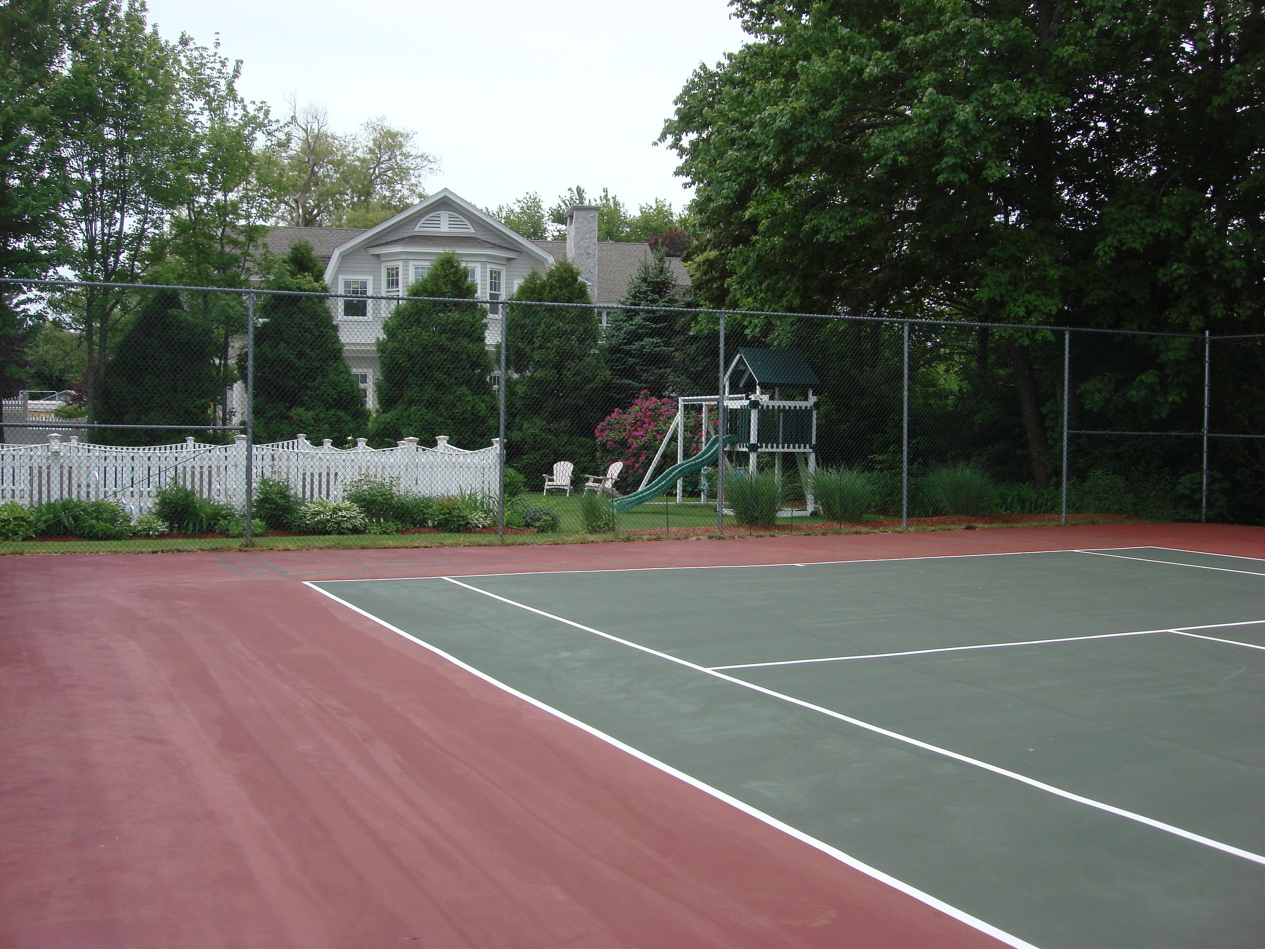 A tennis court with a red surface, surrounded by a white fence and greenery.