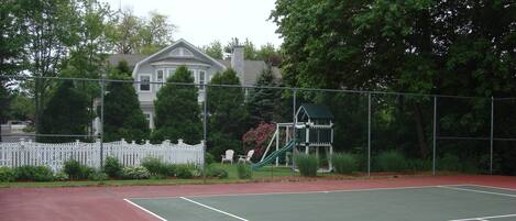 A tennis court with a red surface, surrounded by a white fence and greenery.