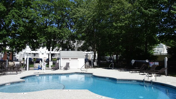 A swimming pool with lounge chairs and a covered area, surrounded by trees and a building in the background. - Pink Blossom Family Resort (Ogunquit)