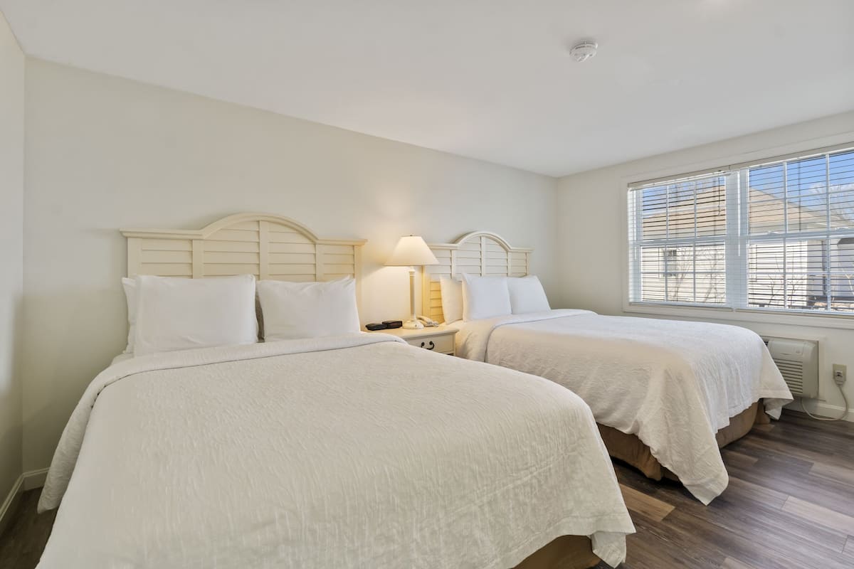 Een hotelkamer met twee bedden, een nachtkastje met een lamp en een raam met zonwering.