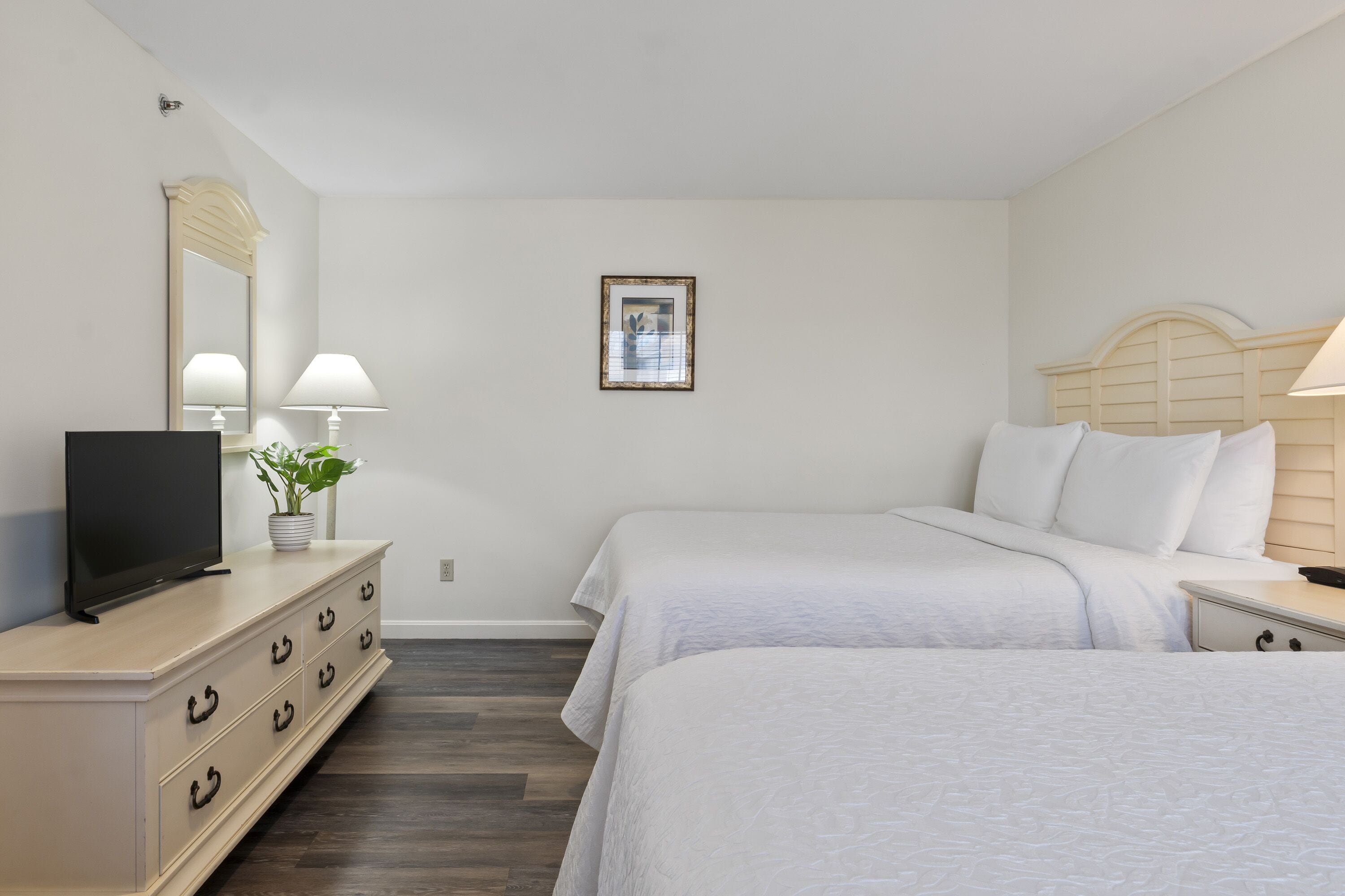 A hotel room with two beds, a TV on a dresser, a mirror, a framed picture on the wall, and a bedside lamp.