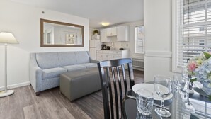 A modern living room with a grey sofa, a black chair, and a dining table set for four. A large mirror hangs on the wall, and a kitchen is visible in the background. - Pink Blossom Family Resort (Ogunquit)