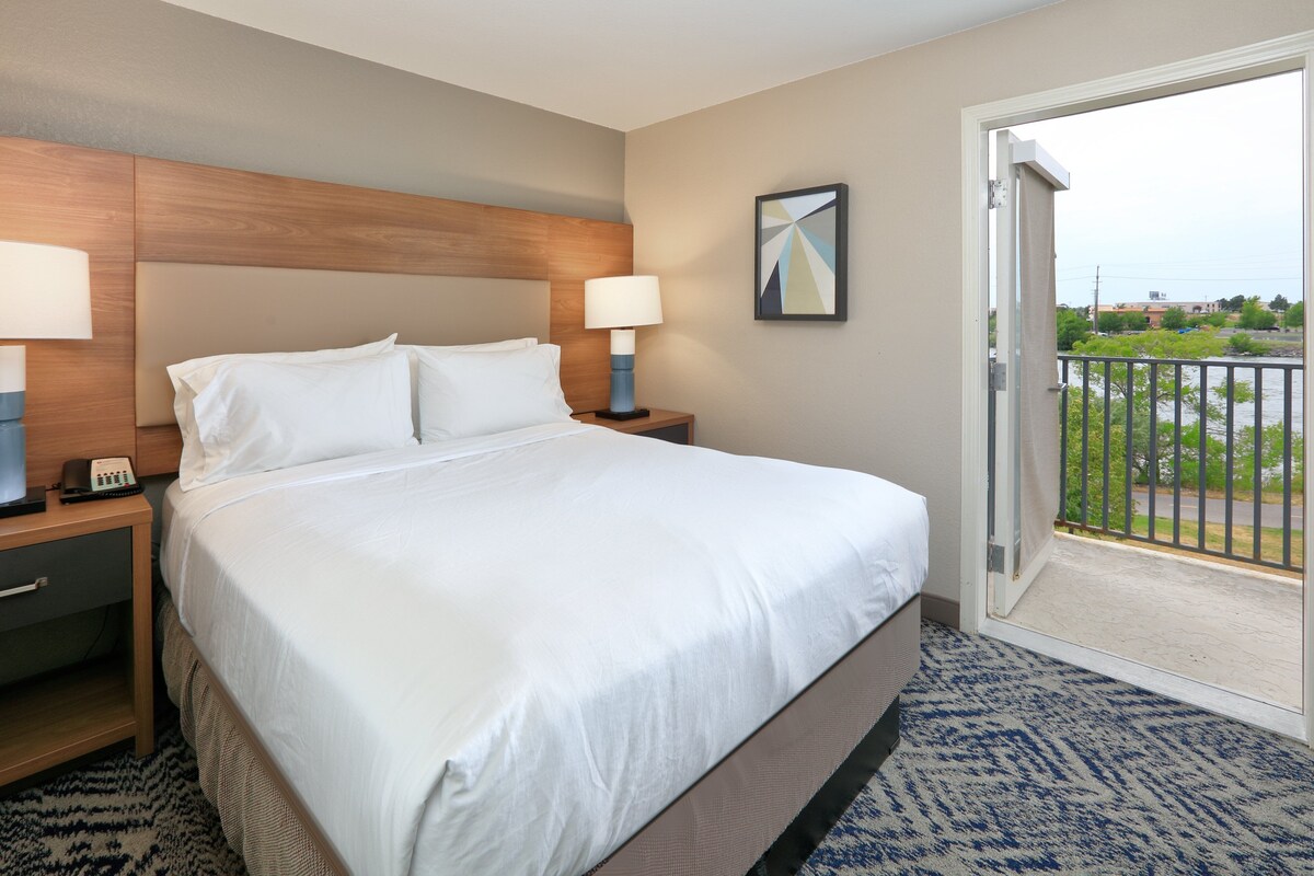 A hotel room with a large bed, two bedside tables with lamps, a nightstand, a door to a balcony, and a framed picture on the wall.