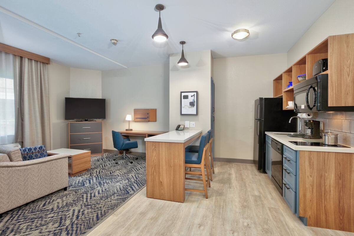 A modern kitchen with wooden cabinets, a microwave, and a sink. There is a dining area with a table and chairs, and a TV in the living room.