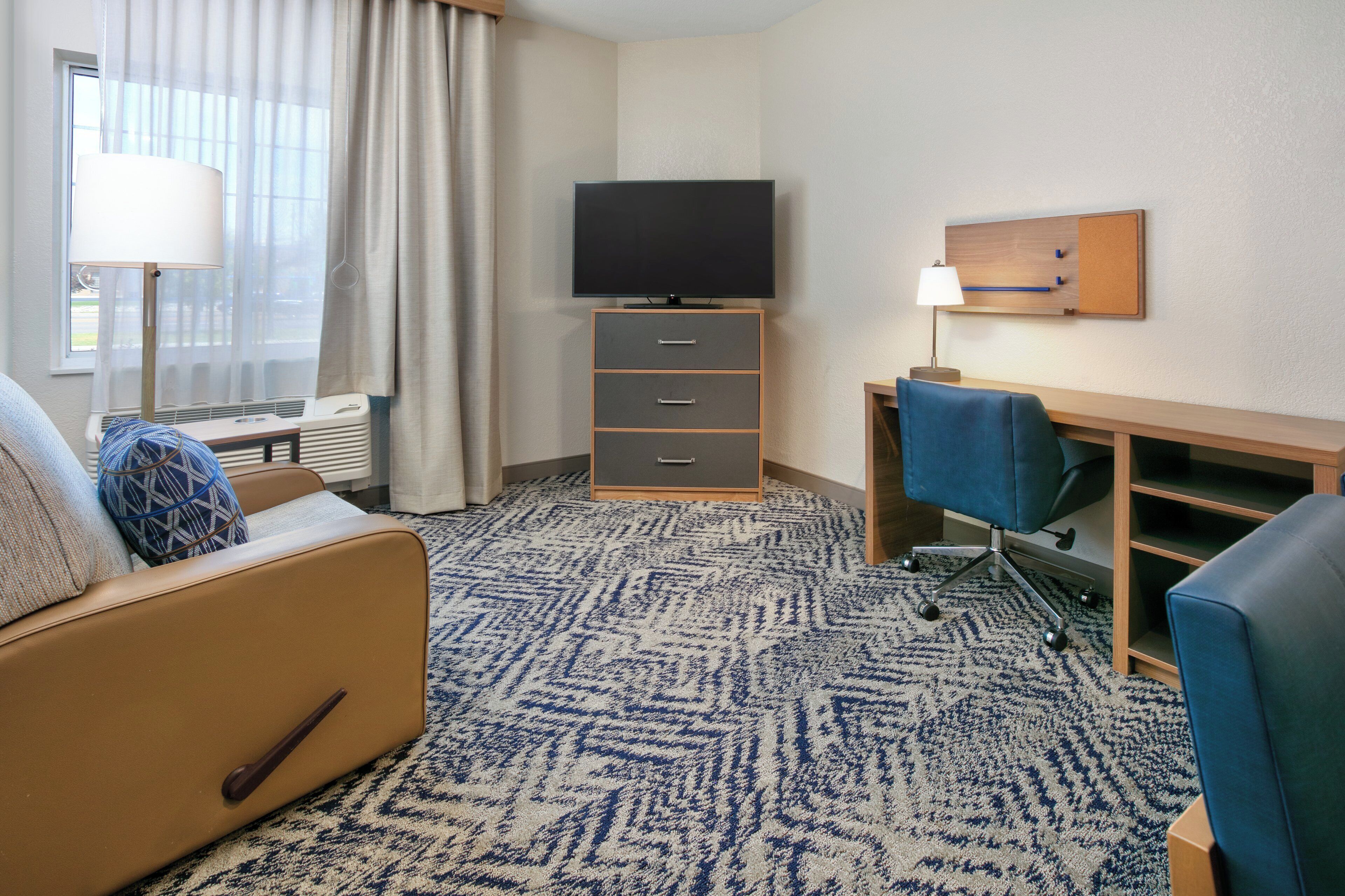 Habitación de hotel con sofá, escritorio con silla, televisión, lámpara y ventana con cortinas.
