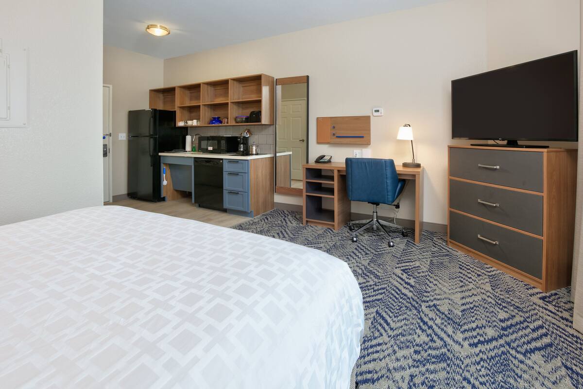 A hotel room with a bed, kitchenette, desk, chair, TV, and wardrobe.