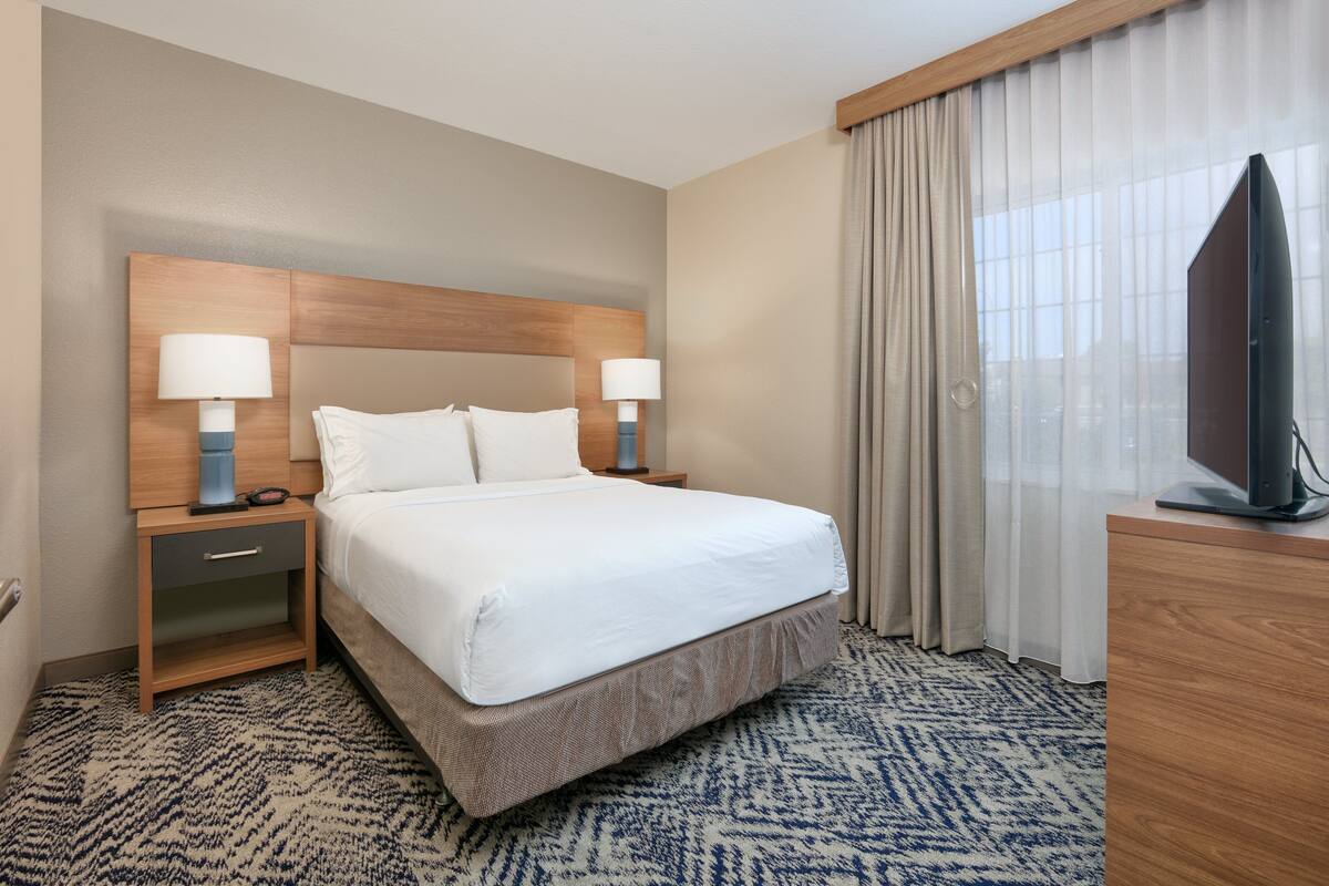 A hotel room with a large bed, two bedside tables with lamps, a television on a wooden cabinet, and a patterned carpet.