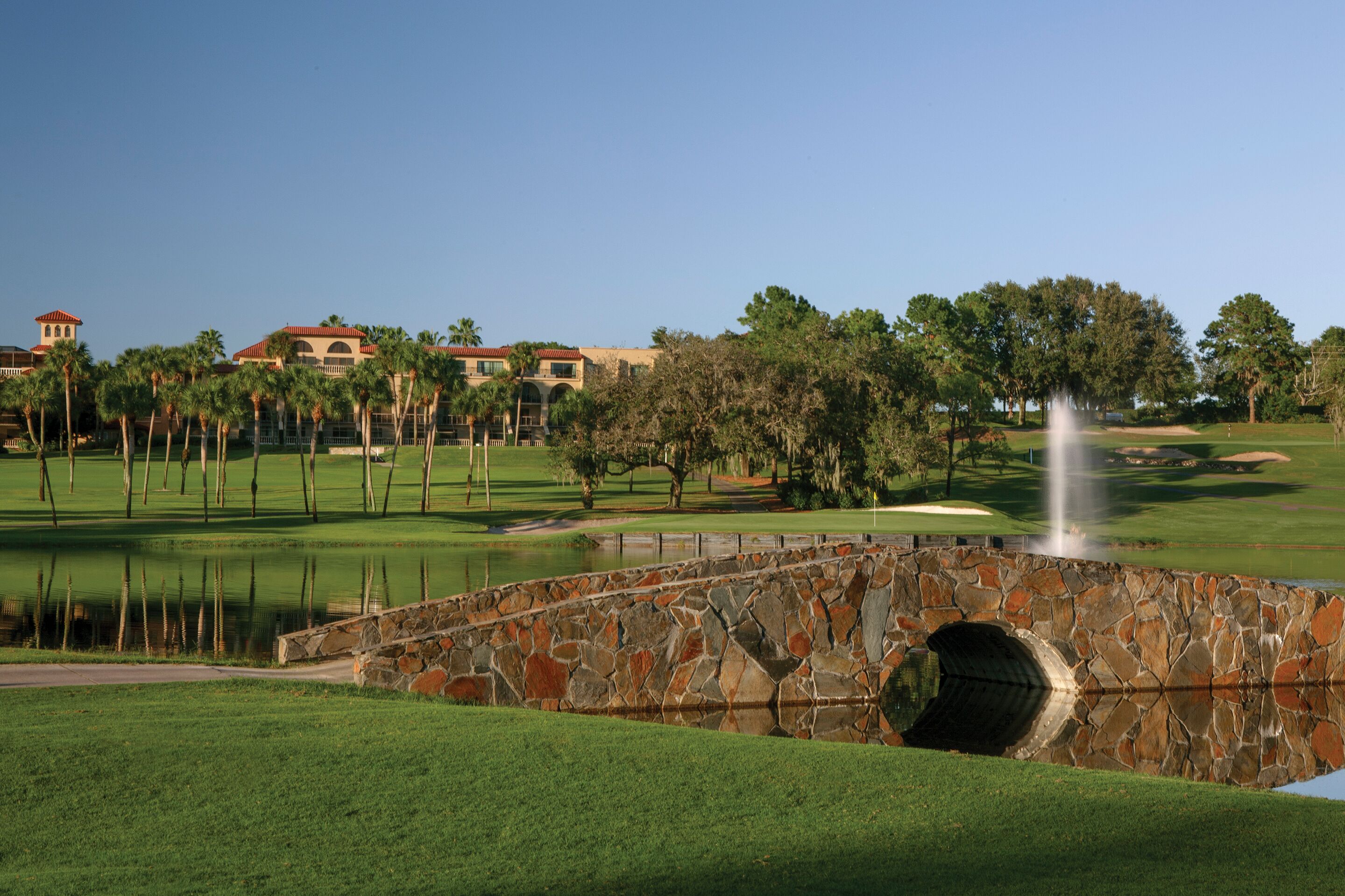 A stone bridge over a pond with a fountain, surrounded by a well-maintained golf course and palm trees.