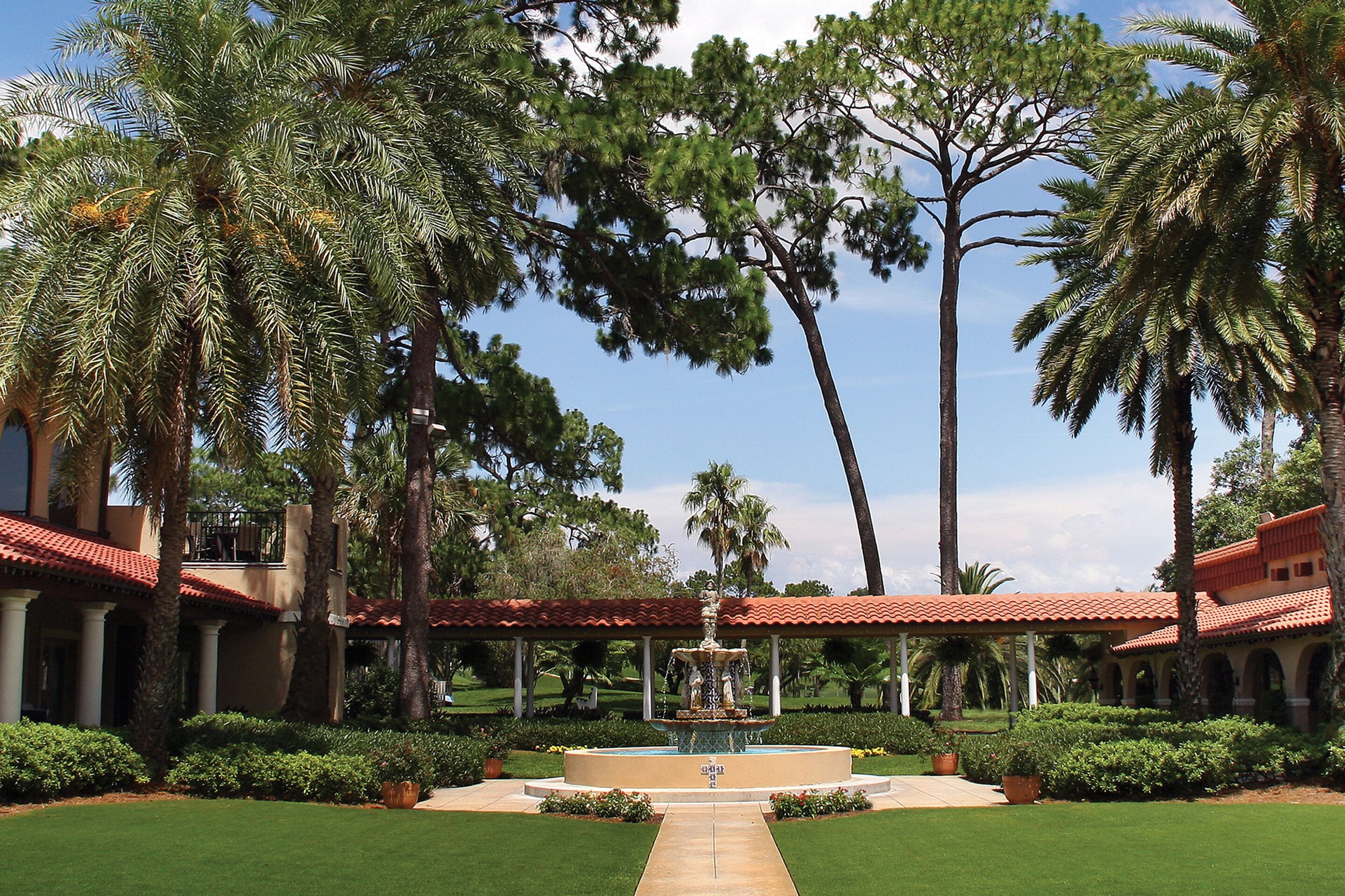 Un patio con una fuente, rodeado de palmeras y un edificio de techo rojo.