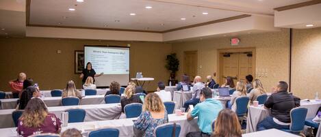 A conference room with rows of tables and chairs, a presentation screen, and attendees seated facing the screen.