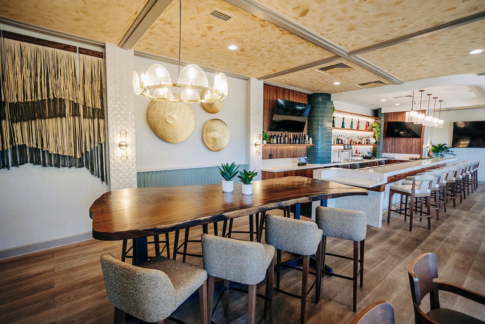 A modern bar area with a long wooden counter, multiple bar stools, and a high ceiling with exposed beams.